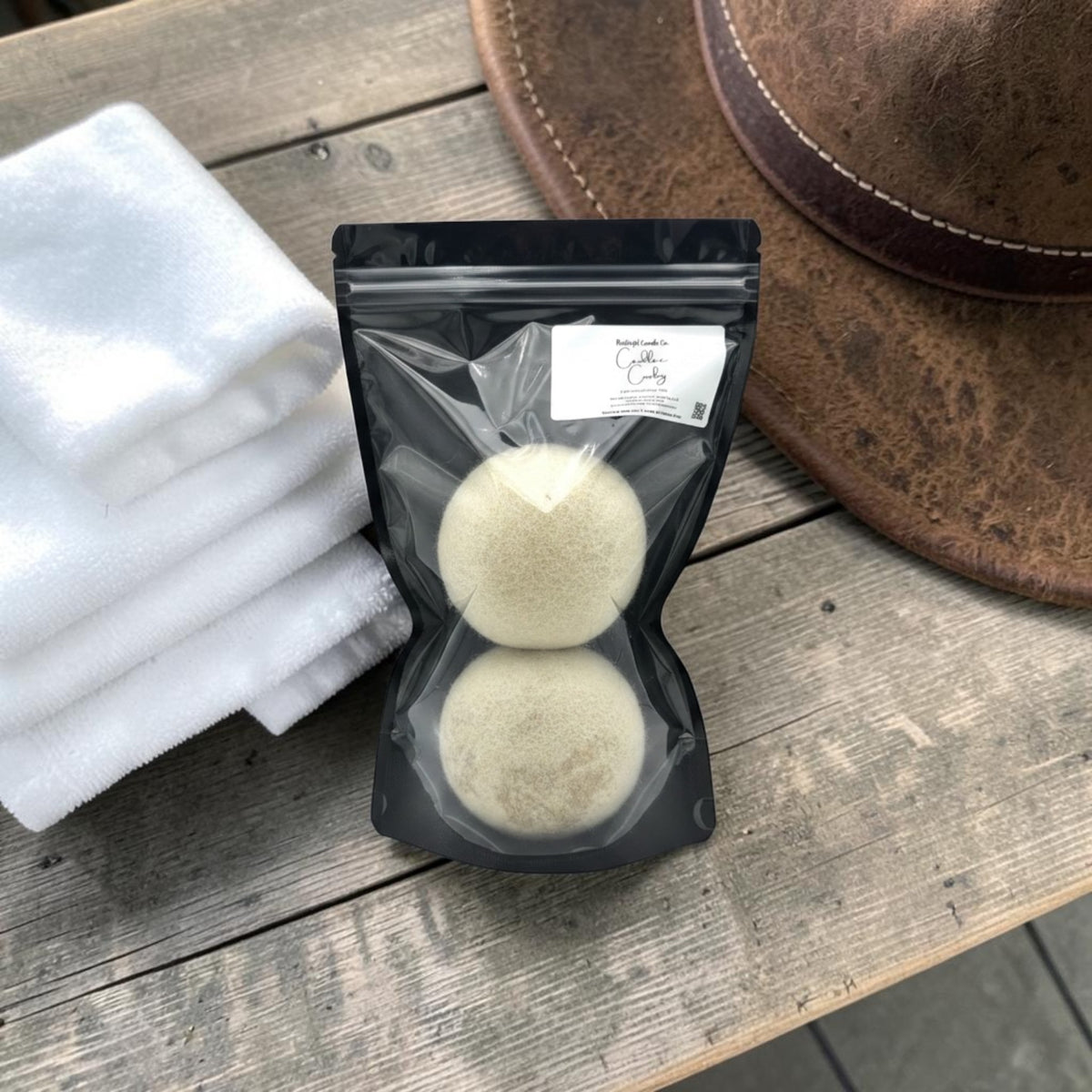 A black resealable pouch containing two round beige exfoliating pads, placed on a rustic wooden surface next to folded white towels and part of a brown hat. A small white label is attached to the pouch.