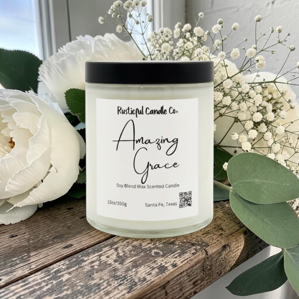 Candle labeled 'Amazing Grace' from Rusticful Candle Co. surrounded by white flowers and green leaves on a wooden surface.