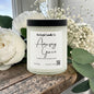 Candle labeled 'Amazing Grace' from Rusticful Candle Co. surrounded by white flowers and green leaves on a wooden surface.