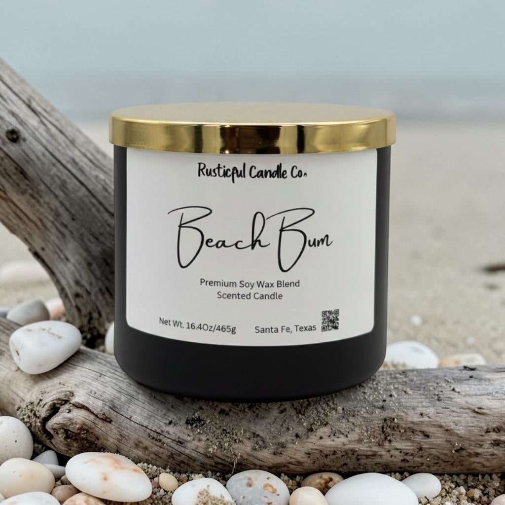Candle labeled 'Beach Bum' from Rusticful Candle Co. on a beach setting with driftwood and pebbles.