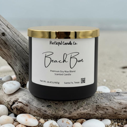 Candle labeled 'Beach Bum' from Rusticful Candle Co. on a beach setting with driftwood and pebbles.