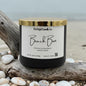 Candle labeled 'Beach Bum' from Rusticful Candle Co. on a beach setting with driftwood and pebbles.