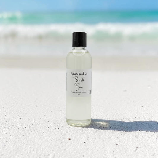 A clear bottle of 'Beach Breeze' fragrance oil by Fragrant Land Co. with a black cap, standing on white sand with a blurred beach and ocean background.