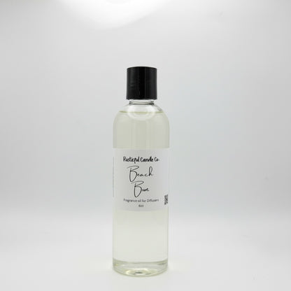 A clear glass bottle of Beach Breeze Fragrance Oil by Rustical Candle Co., featuring a white label and a black screw cap, set against a plain white background.