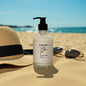 Bottle of body lotion with a pump on sand, straw hat, and sunglasses in the background