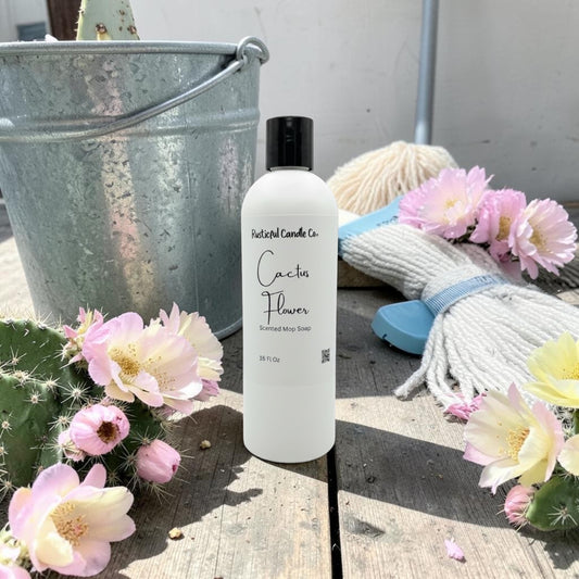 A frosted white bottle of Natural Candle Co. Cactus Flower scented soy wax candle stands on a rustic wooden surface, surrounded by pink cactus flowers, a galvanized metal bucket, and a white rope with a blue tie.