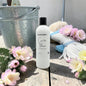 A frosted white bottle of Natural Candle Co. Cactus Flower scented soy wax candle stands on a rustic wooden surface, surrounded by pink cactus flowers, a galvanized metal bucket, and a white rope with a blue tie.