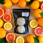 Two bath bombs in a black packaging surrounded by a variety of citrus fruits on a wooden surface.
