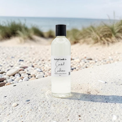 A bottle of daylight candles 'Candle Cushion' scented oil diffuser and candle, placed on a sandy beach with pebbles and dune grass in the background.