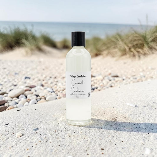 A bottle of daylight candles 'Candle Cushion' scented oil diffuser and candle, placed on a sandy beach with pebbles and dune grass in the background.