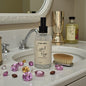 Body mist bottle labeled 'Crystal Rouge' on a bathroom counter with decorative stones and a mirror in the background.