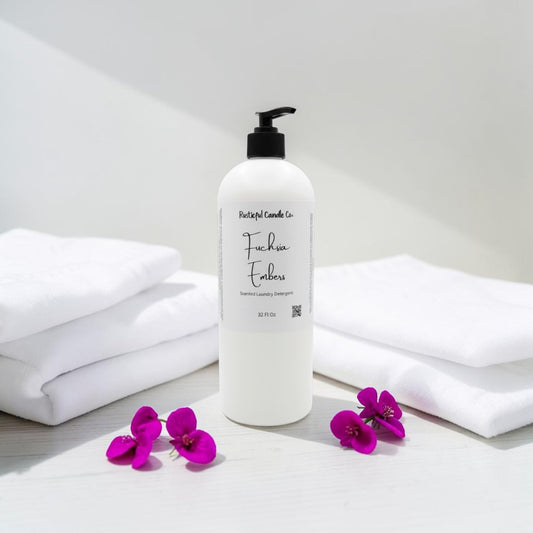 A white pump bottle of Fuchsia Embers laundry detergent by Fungal Candles Co, styled on a white surface with folded white towels and purple flowers.
