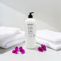 A white pump bottle of Fuchsia Embers laundry detergent by Fungal Candles Co, styled on a white surface with folded white towels and purple flowers.