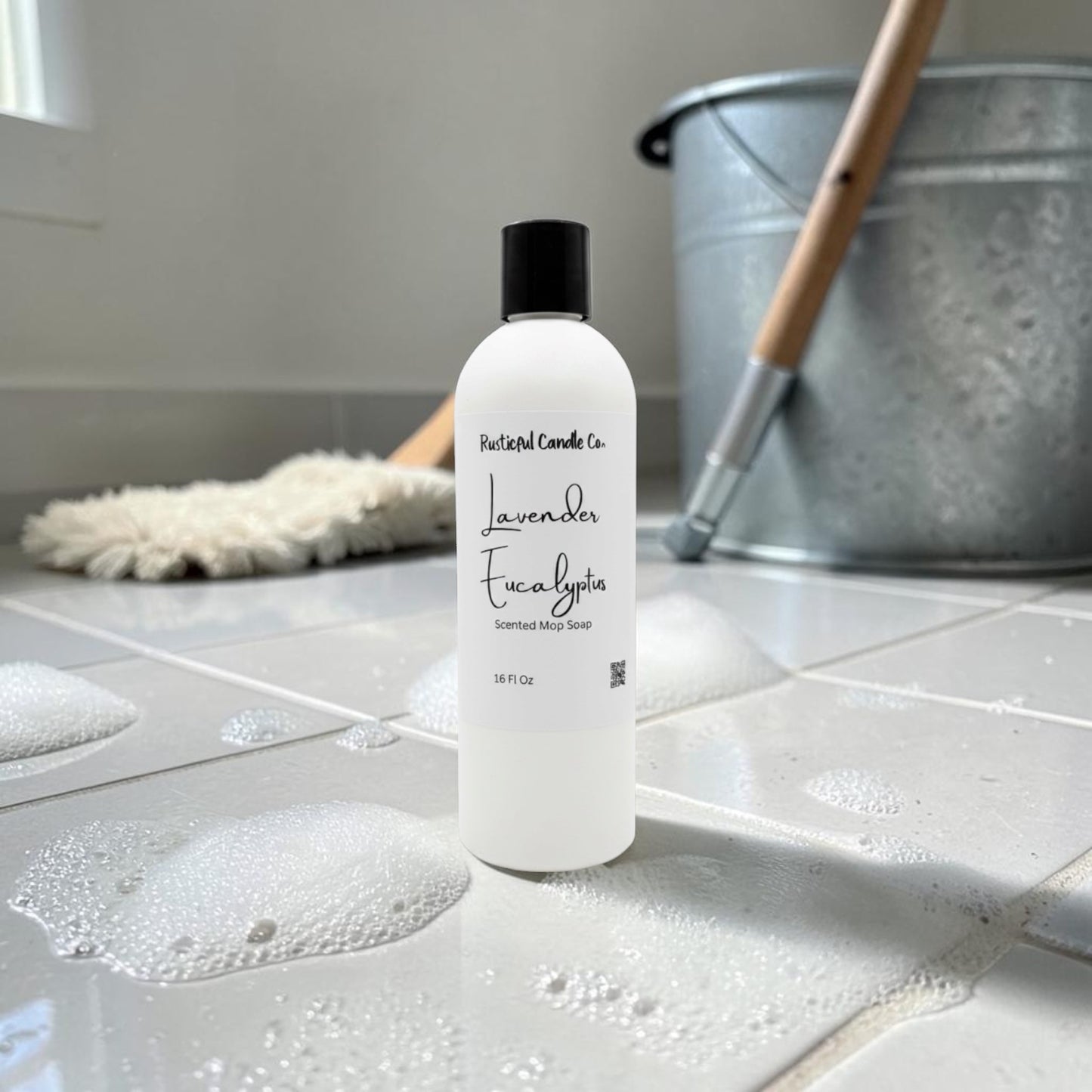 A bottle of Natural Candles Co. Lavender Eucalyptus Treated Wood Cleanser stands on a tiled bathroom floor with soap bubbles, a mop, and a metal bucket in the background.