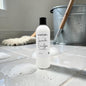 A bottle of Natural Candles Co. Lavender Eucalyptus Treated Wood Cleanser stands on a tiled bathroom floor with soap bubbles, a mop, and a metal bucket in the background.