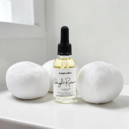 A small glass dropper bottle labeled 'Moonlight Romance' by Pretty Girl Candle Co, filled with pale yellow liquid, standing on a white surface between two white bath bombs against a bright white background.