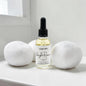 A small glass dropper bottle labeled 'Moonlight Romance' by Pretty Girl Candle Co, filled with pale yellow liquid, standing on a white surface between two white bath bombs against a bright white background.