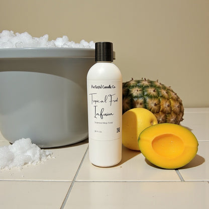 Bottle of 'Tropical Fruit Infusion' soap next to a pineapple and mango on a tiled surface.