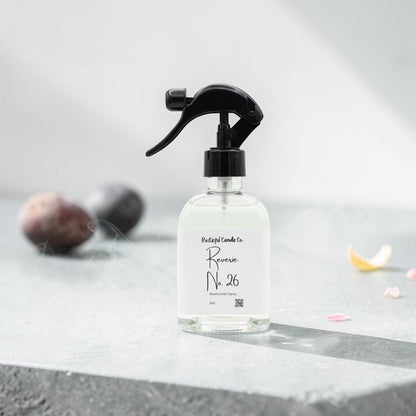 A clear glass spray bottle with a black trigger sprayer and a white label reading 'Botanicals Lane Reverse No. 26', set on a gray stone surface with soft-focus decorative stones in the background.