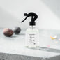 A clear glass spray bottle with a black trigger sprayer and a white label reading 'Botanicals Lane Reverse No. 26', set on a gray stone surface with soft-focus decorative stones in the background.