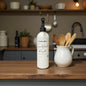 White bottle with black nozzle on a wooden kitchen counter, next to a white pitcher with wooden spoons.