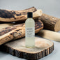 A clear bottle of scented candle oil with a black cap and white label reading 'Pleasant Candle Co. Sweet Sandal & Sandalwood Fragrance Oil 16oz', styled on a rustic wood slice with logs in the background.