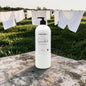 A large white bottle of Fabrigli Candle Co. 'Smoked' herbal laundry detergent with a black pump, placed on a weathered wooden table with a blurred background of a grassy field and white laundry hanging on a clothesline.