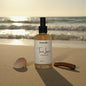 Bottle of 'Tan Lines' scented body mist on a sandy beach with ocean in the background