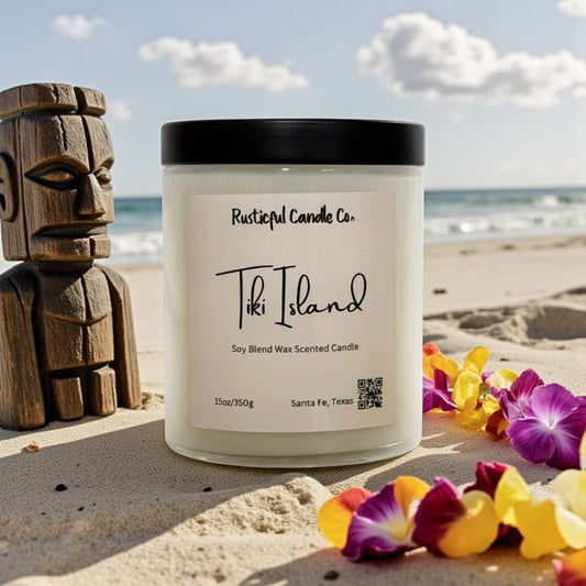 Candle labeled 'Tiki Island' from Rusticful Candle Co. on a sandy beach with flowers and a wooden tiki figure.
