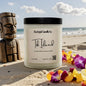 Candle labeled 'Tiki Island' from Rusticful Candle Co. on a sandy beach with flowers and a wooden tiki figure.
