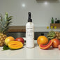 White bottle labeled 'Tropical Fruit Infusion' surrounded by pineapples, mangoes, and papayas on a kitchen counter.