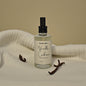 Bottle of Vanilla Cashmere scented body mist with vanilla beans on a beige background