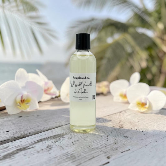 A clear bottle of fragrance oil with a black cap and white label reading 'Paisley Candle Co. Moped Vanilla & Amber' stands on a weathered white wooden surface outdoors, with orchids and blurred palm leaves in the background.
