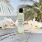 A clear bottle of fragrance oil with a black cap and white label reading 'Paisley Candle Co. Moped Vanilla & Amber' stands on a weathered white wooden surface outdoors, with orchids and blurred palm leaves in the background.