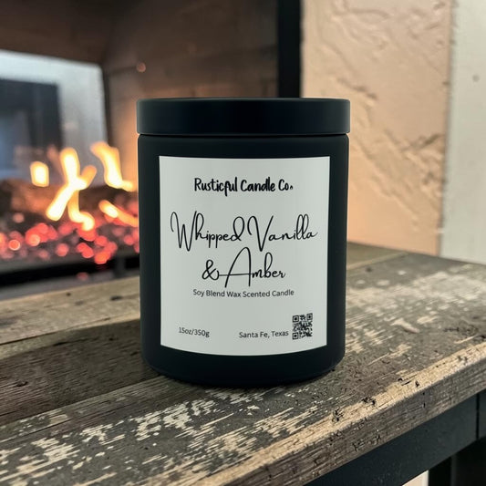 A black candle jar with a white label reading 'Rusticful Candle Co. Whipped Vanilla & Amber' sits on a rustic wooden surface in front of a blurred fireplace.