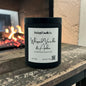 A black candle jar with a white label reading 'Rusticful Candle Co. Whipped Vanilla & Amber' sits on a rustic wooden surface in front of a blurred fireplace.